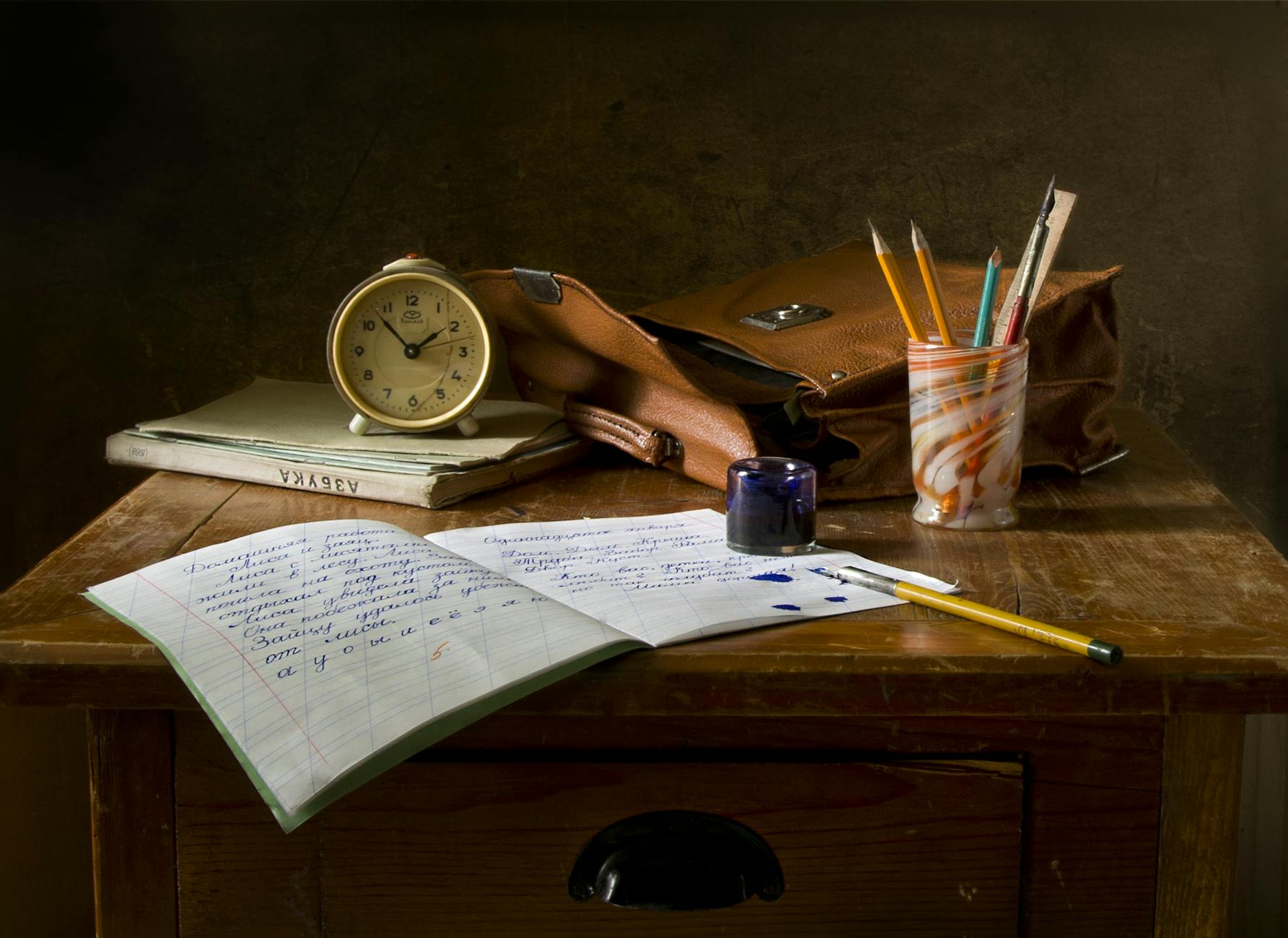 A wooden desk with a notebook, an alarm clock, a brown leather bag, and a container of colored pencils and pens.